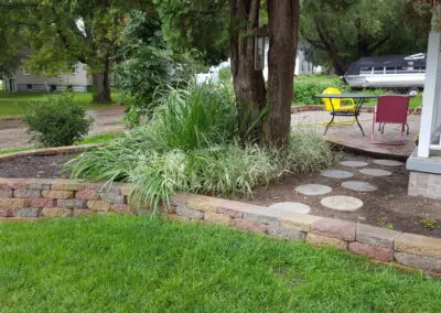 Front yard raised stone garden bed curving around a group of trees with ornamental grasses, circular concrete stepping stones leading to a small brick patio with a yellow metal chair and pink lawn chair beside a porch and parked boat in the background