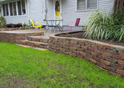 Curved multi-level retaining wall and raised patio made of interlocking red, brown and gray stone blocks in front of a gray vinyl-sided house with a yellow front door, patio table, yellow and pink metal chairs, garden bed with ornamental grasses and freshly seeded lawn