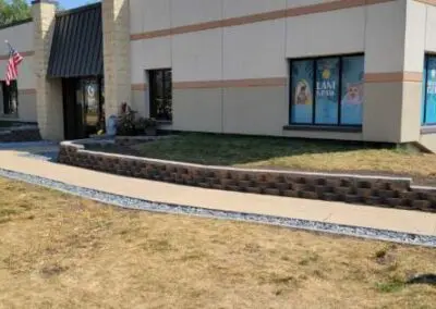 Commercial building exterior with a concrete sidewalk bordered by decorative stone retaining walls and white gravel, a small landscaped entrance with potted plants, American flag by the doorway, and storefront windows displaying pet-themed posters.