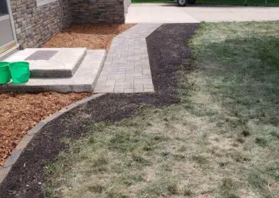 Front yard view of a stone-faced house with concrete steps and a newly installed paver walkway leading to the driveway, mulch beds at the house foundation, bare patchy lawn in foreground, and a covered boat on a trailer parked on the driveway.