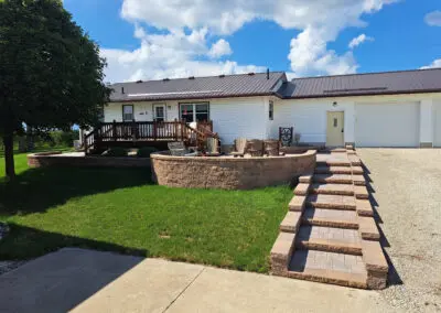 Raised stone patio and terraced paver steps leading to a white ranch home with attached garage, cedar deck and Adirondack chairs under a blue sky, Sharon patio landscaping project