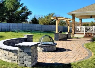 Backyard patio with curved low stone retaining wall and circular stone fire pit with glass dome, paved brick patio leading to a wooden pergola and covered porch with stone columns, lawn and wooden privacy fence in background