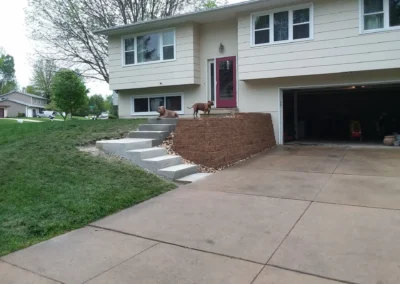 Suburban split-level house with a raised brick retaining wall and concrete steps leading from driveway to a red front door; two dogs rest on the top of the steps and a two-car garage is open to the right, with lawn and trees visible on the left.