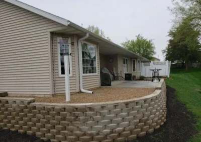 Backyard patio with concrete paver retaining wall curving around a raised gravel bed, beige vinyl-sided ranch house with sliding glass door, downspout, covered grill and patio furniture under an eave, green lawn and trees in background