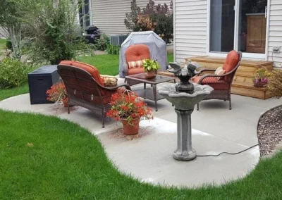 Backyard concrete patio with curved edges featuring wicker seating with orange cushions around a small coffee table, potted red flowering plants, a concrete birdbath with an electrical cord, a covered grill, wooden steps to a sliding glass door, and surrounding green lawn and landscaping