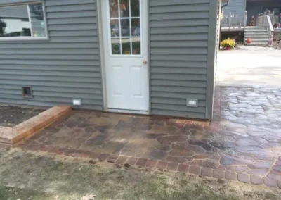 Stamped concrete patio and walkway in brown tones leading to a white back door of a gray-sided house with a small raised planting bed at left and adjoining stamped path toward a raised deck area in the background