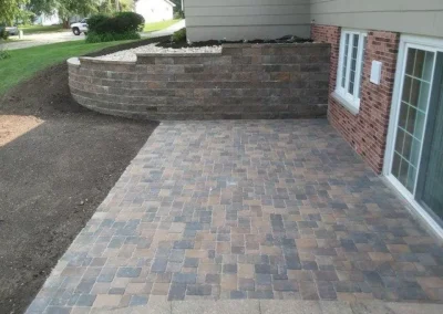 Patio with interlocking gray and tan pavers leading from a brick house with sliding glass door to a raised curved retaining wall filled with white gravel and mulch, lawn and suburban street visible beyond