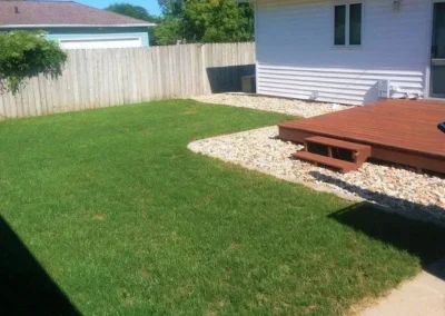 Backyard lawn with a small wooden deck and steps beside a white house, light-colored rock border separating the deck from the green grass, wooden privacy fence and neighboring garage in background