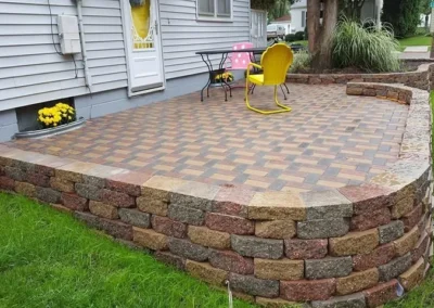 Small backyard raised patio with interlocking multicolored pavers and a curved stacked stone retaining wall, featuring a yellow metal chair, small round table with pink polka-dot cushion, a house door and planter of yellow flowers against a lawn
