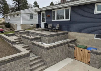 Front yard showing multi-level gray block retaining walls and stepped patio with gravel-filled planters, concrete stairways leading to a small porch with two chairs at a blue-sided single-story house, with a wooden gate concealing green and blue recycling bins.
