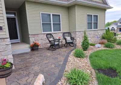Front porch with stamped concrete walkway edged by river rock and curved lawn, two wicker chairs and small table with potted flowers against a beige vinyl-sided house with stone veneer and double windows, small dog walking on the path