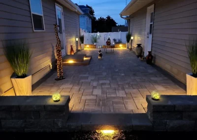 Evening backyard patio with paver flooring, low built-in stone seating walls and recessed warm LED step lights, decorative planters and vases along both house walls, a small wooden table and lanterns near a white privacy fence, and two small dogs sitting on the patio under a twilight sky