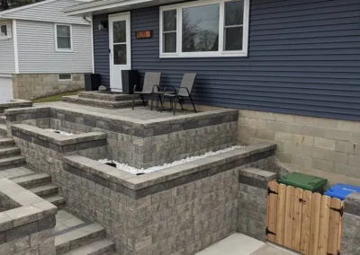Front yard of a single-story blue-sided house with a raised multi-level gray concrete block retaining wall and patio, concrete steps leading up to a small furnished entry patio with two chairs, decorative white stone in planter ledges, house number plaque by the white door, and a small wooden hinged gate beside green and blue recycling bins.