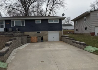 Driveway leading to a single-car garage set into a raised split-level house with dark blue siding; concrete retaining walls with caps flank the wide concrete driveway, small wooden gate and potted evergreen beside the garage, overcast sky and bare winter trees in background