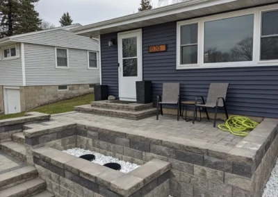 Front porch patio with gray interlocking pavers and tiered retaining walls, two outdoor chairs and small table beside a white storm door and house number 1020 on navy blue siding, decorative white stones in a lower planter and coiled green garden hose on the right