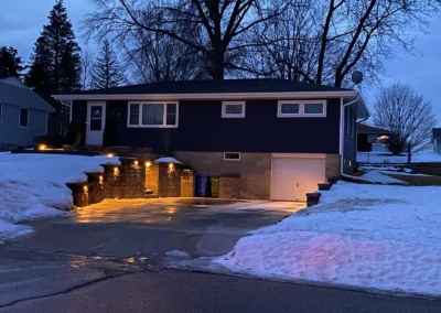 Evening suburban split-level house with lit retaining-wall and driveway lights, snow-covered yard and a closed single-car garage