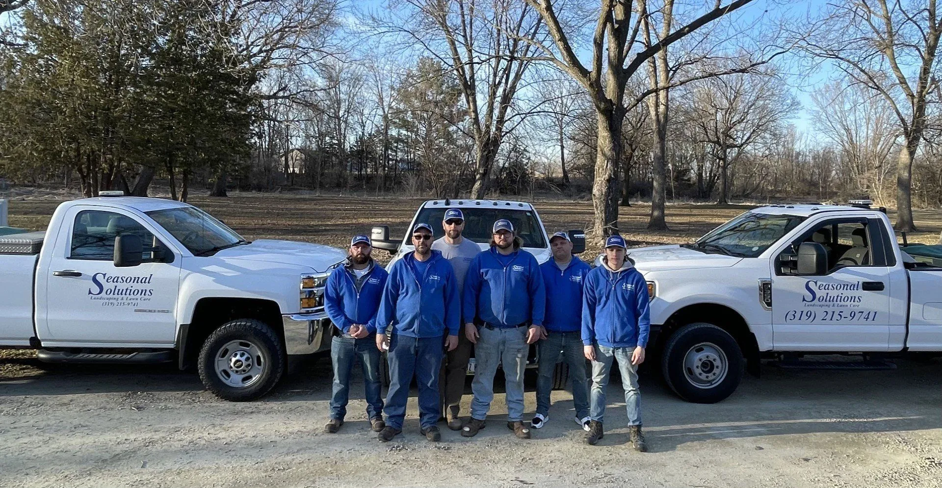 Seasonal Solutions landscaping crew in blue jackets standing in front of three white company trucks with Seasonal Solutions logos and phone number, parked on a rural lot with bare trees and winter grass